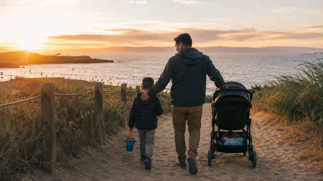 A dad and his young son walk side by side down a sandy path toward the sea at sunset
