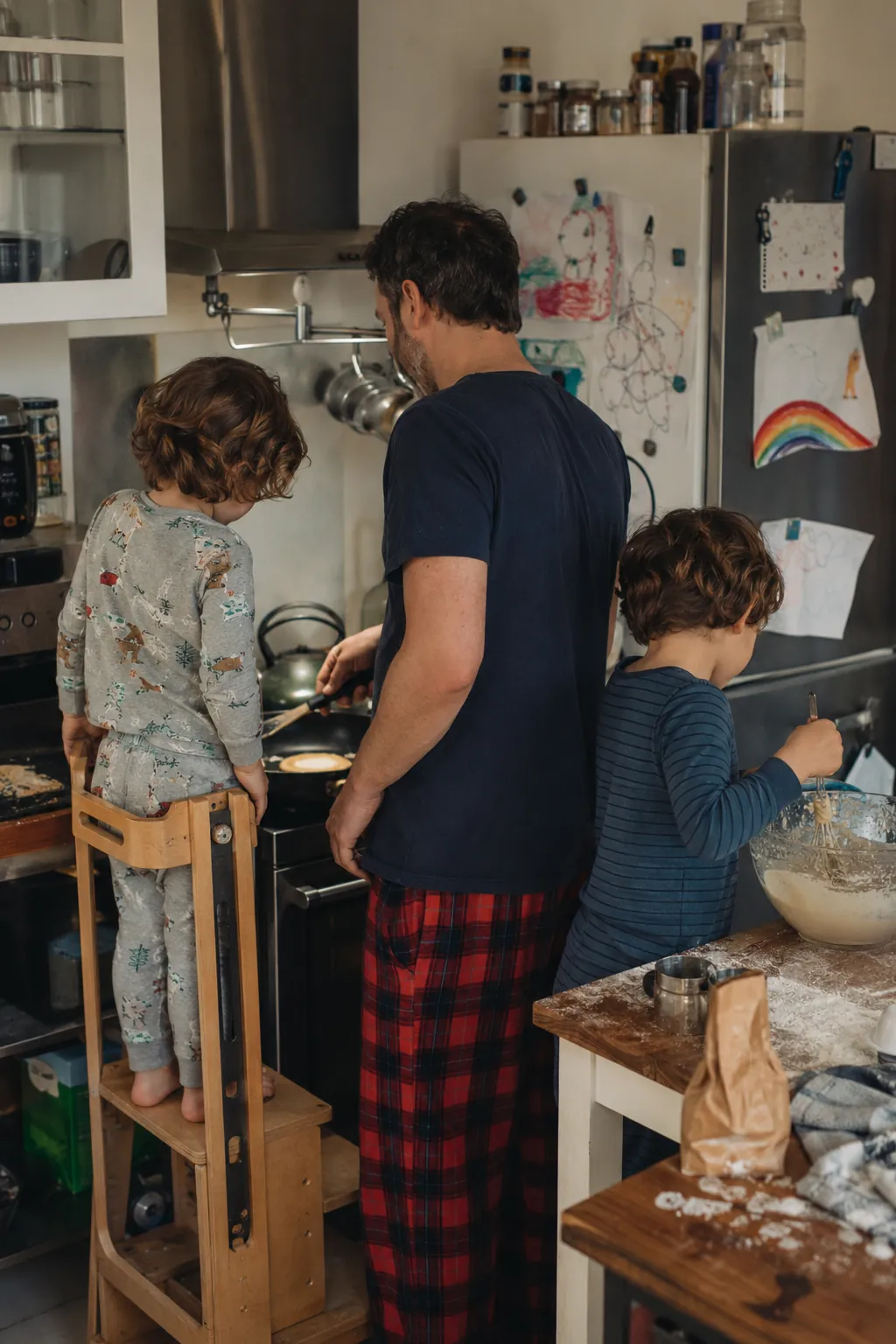 Two kids in pajamas cook side by side with their dad, one standing on a step stool