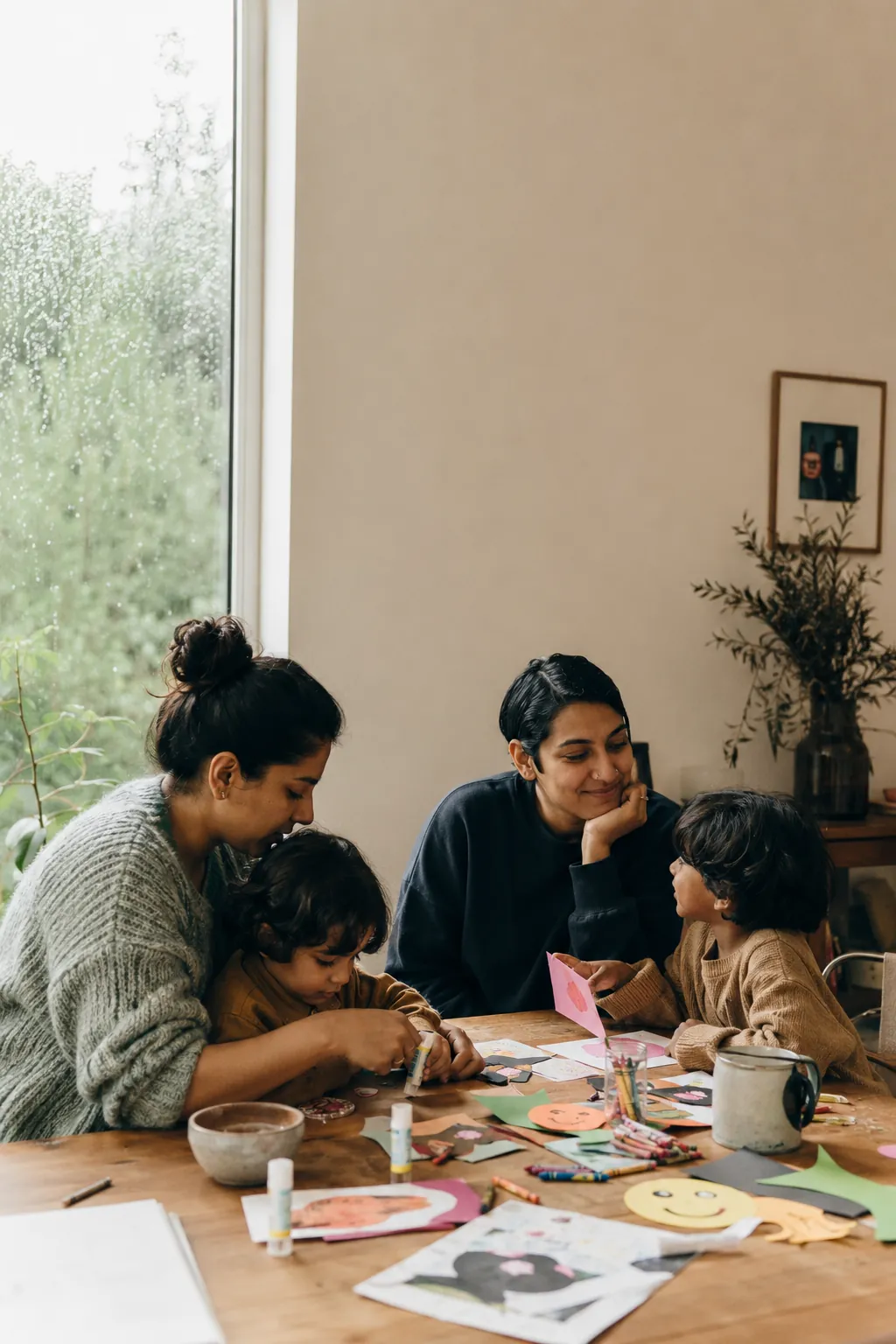 Two moms help two kids glue felt shapes and paper decorations at a craft table covered in crayons and glue sticks