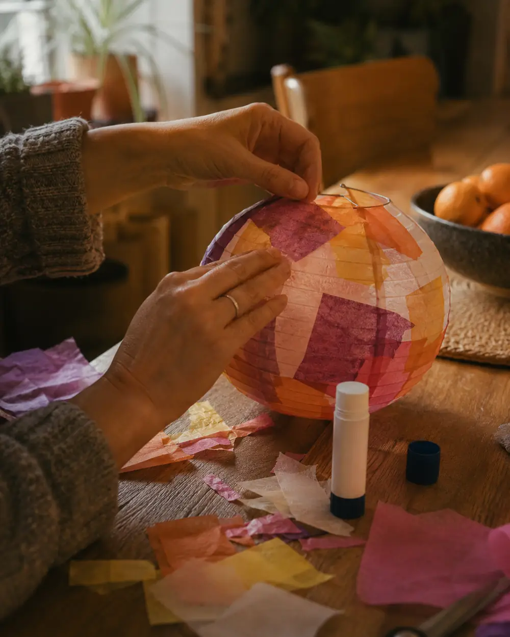 Hands folding and gluing strips of colored paper into a lantern on a table