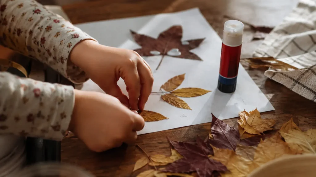 A small child's hands placing dried autumn leaves onto a sheet of paper beside a glue stick