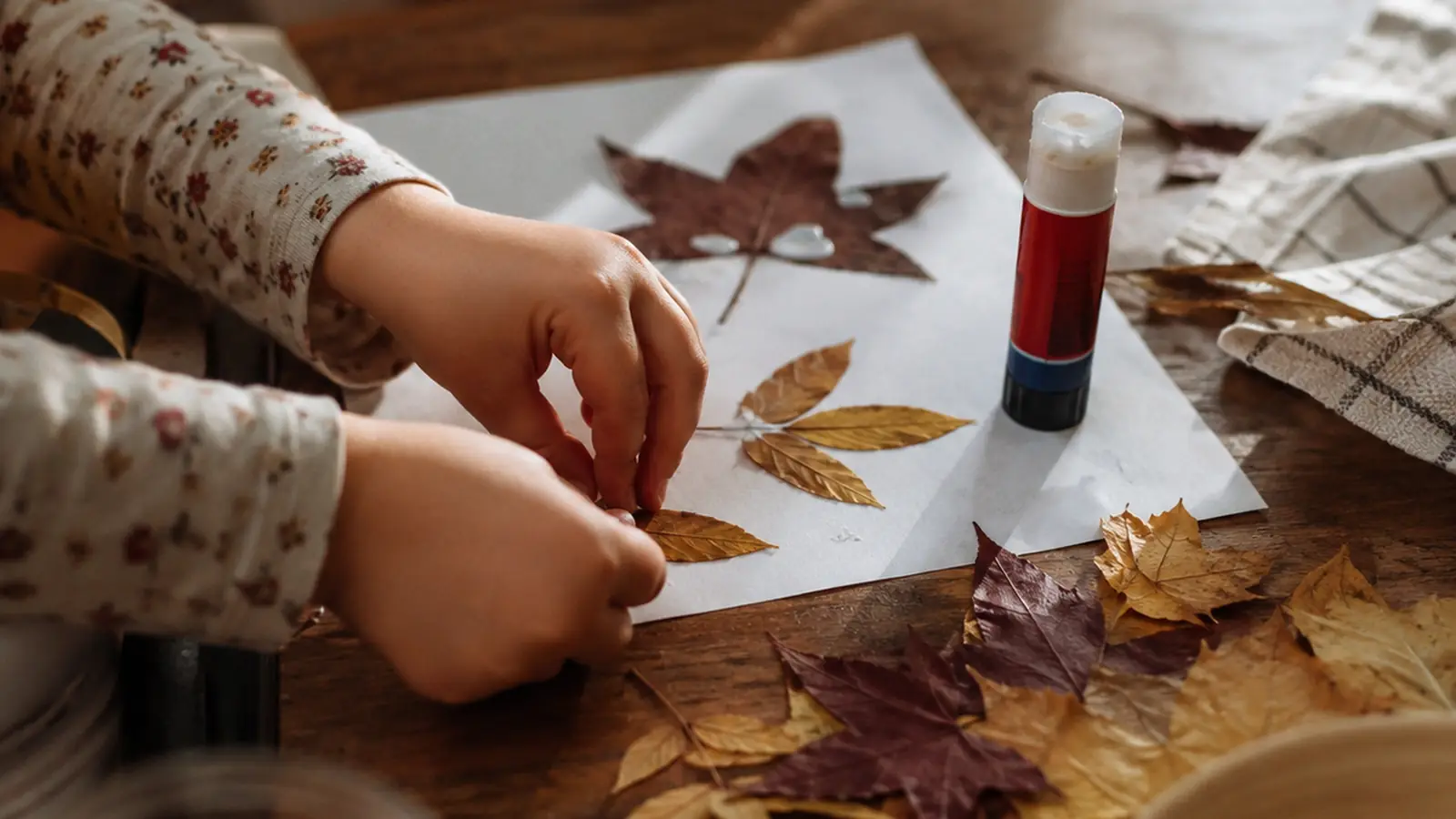 A small child's hands placing dried autumn leaves onto a sheet of paper beside a glue stick
