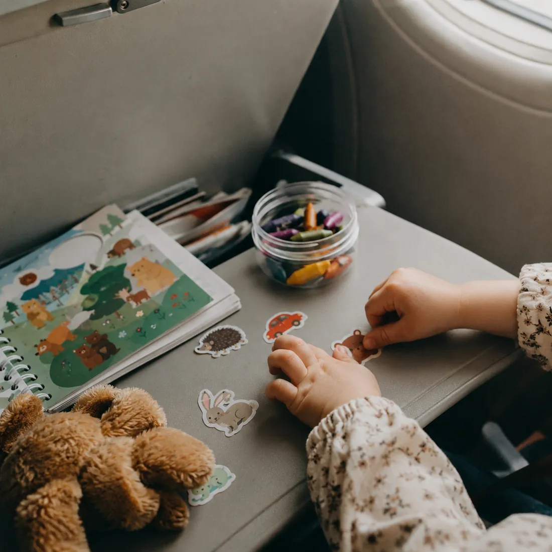 A toddler plays with stickers, crayons and a small stuffed animal spread across an airplane tray table