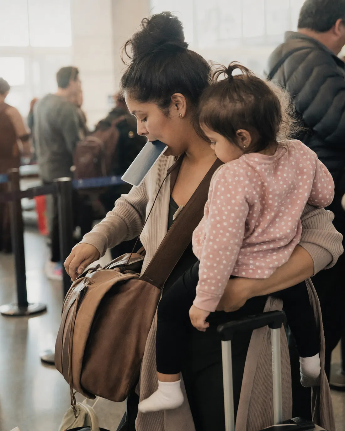 A parent holds a toddler on one hip while carrying a boarding pass in their teeth and pulling a rolling bag through the TSA security line