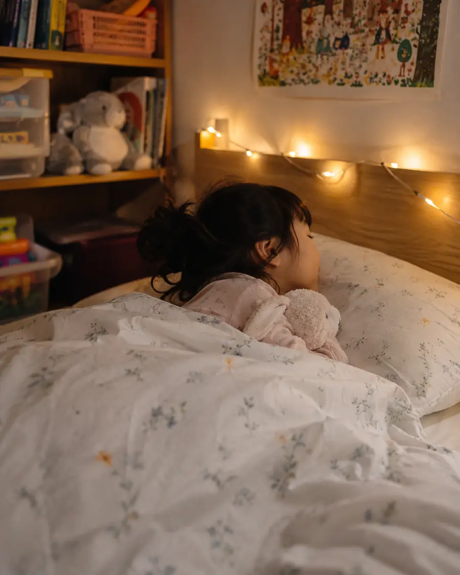 A young girl falls asleep in bed under warm fairy lights, holding a soft toy