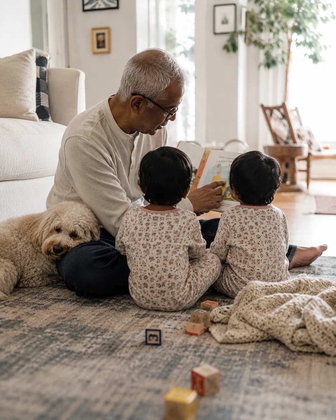 Grandfather reads a picture book to twin toddlers on the floor with their dog beside them