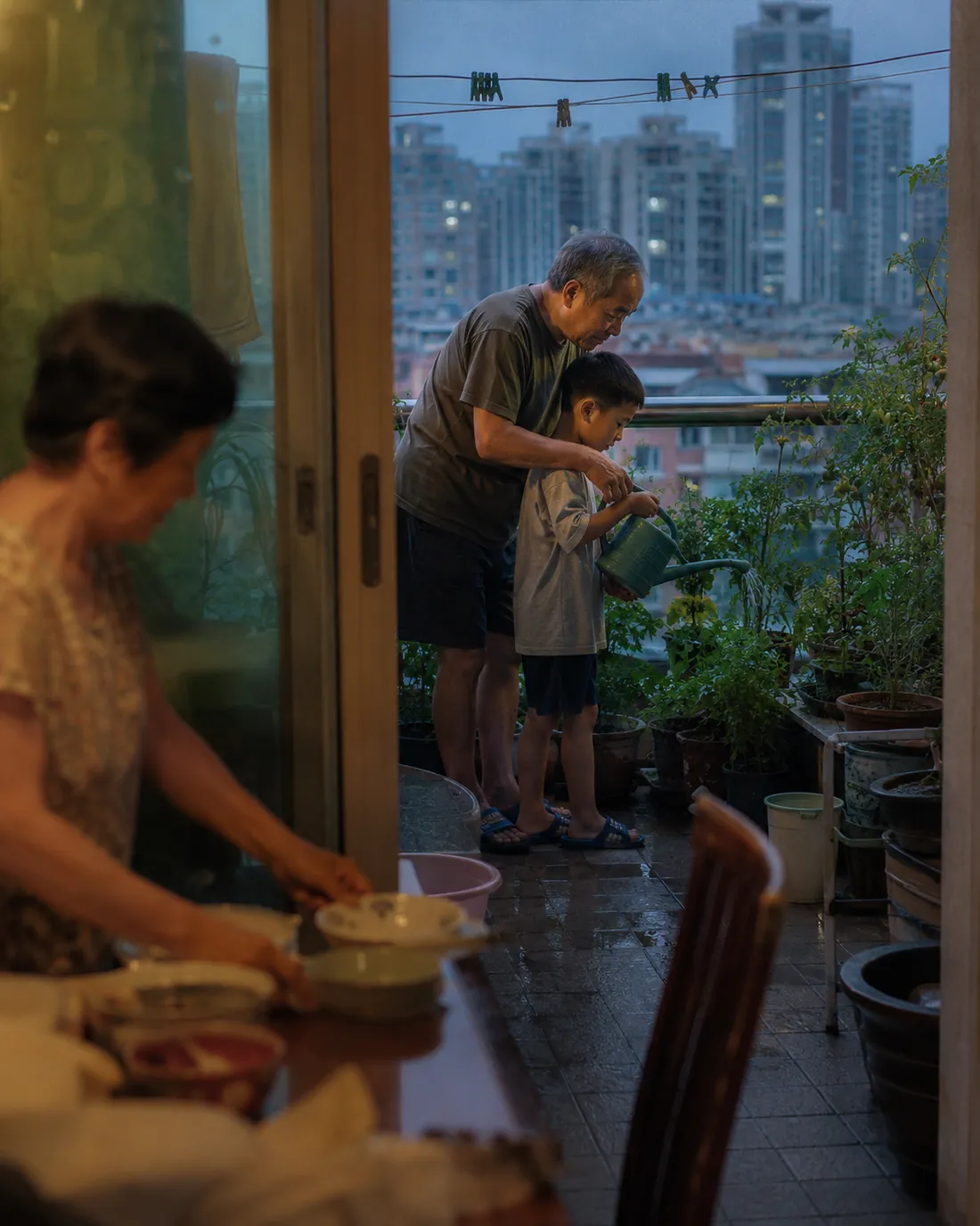 A grandfather and grandson water plants on a balcony at dusk