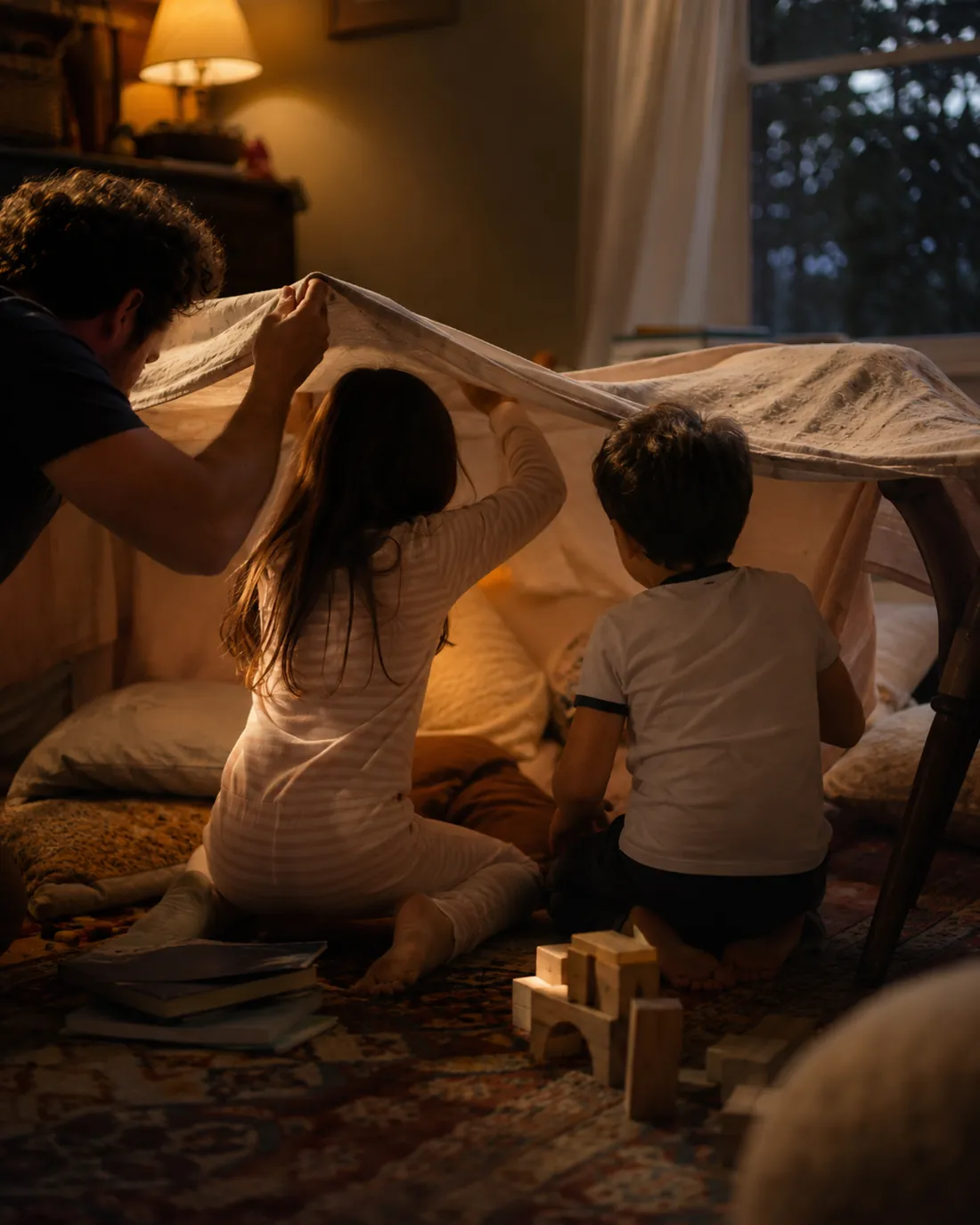 Two children help build a blanket fort in a warmly lit living room in the evening