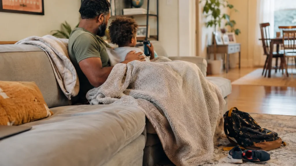 A father and young child sit under a blanket on the couch watching a movie together after school