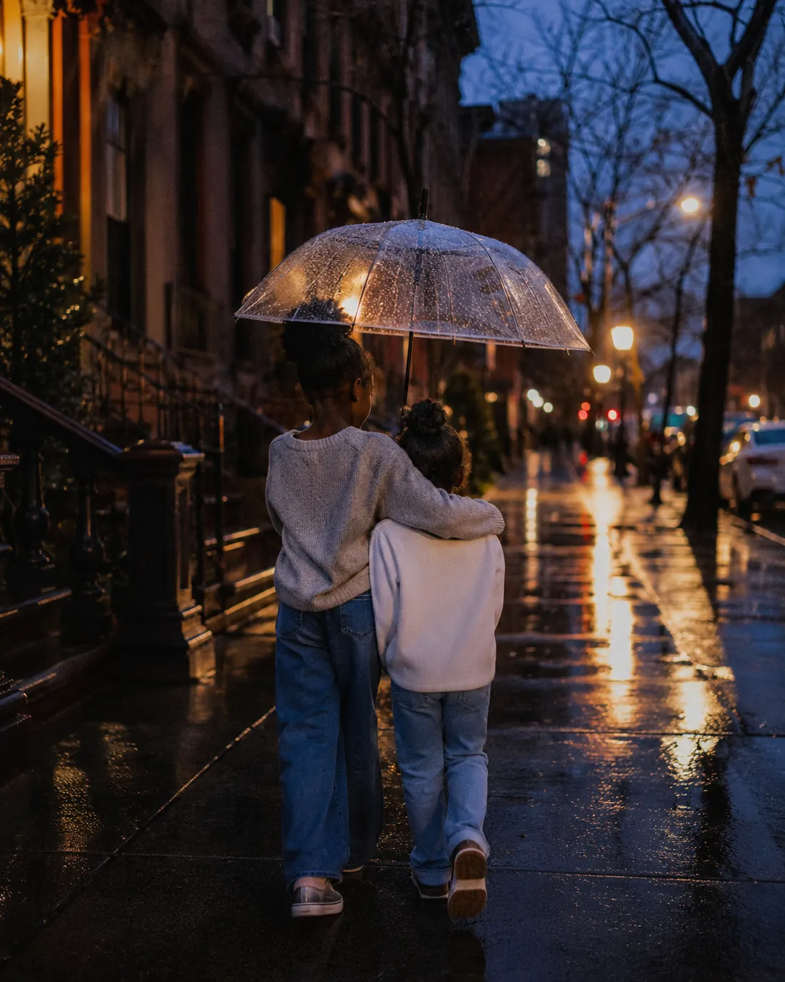 Two kids in pale sweaters walk under a clear umbrella down a dark, rain-wet city street lined with streetlights