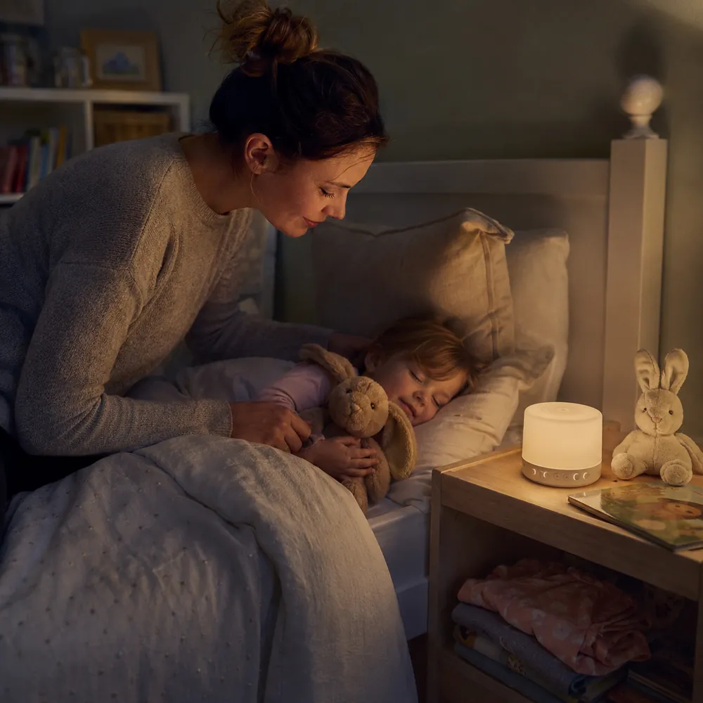 A mother leans over a sleeping toddler hugging a stuffed bunny, with a glowing night light on the bedside table