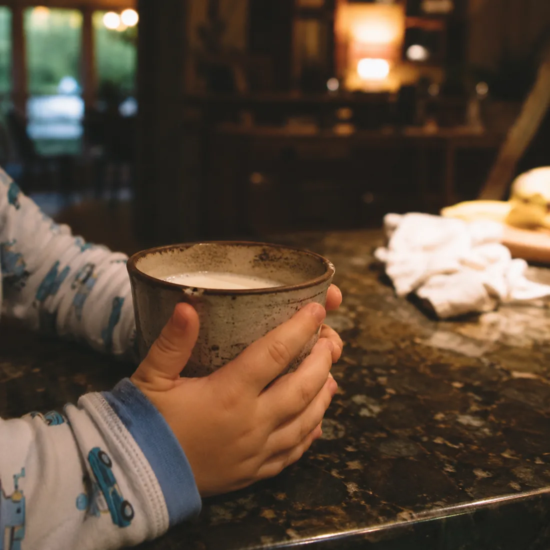A child in pyjamas holding a mug of warm milk with both hands on a kitchen counter in the evening