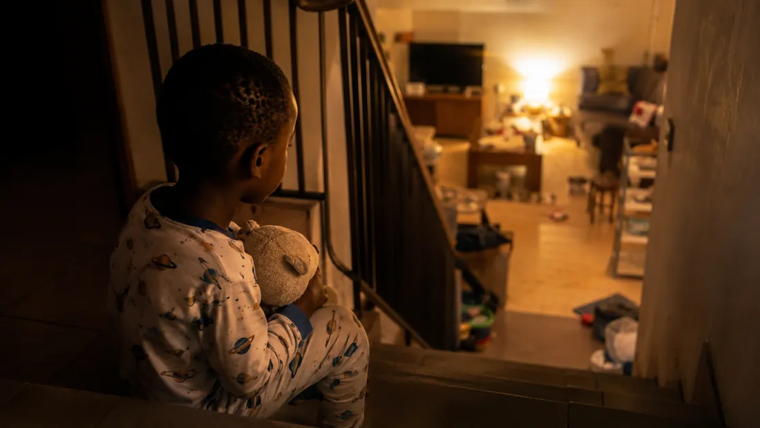 A child in pajamas sits on the stairs late in the evening, holding a stuffed toy