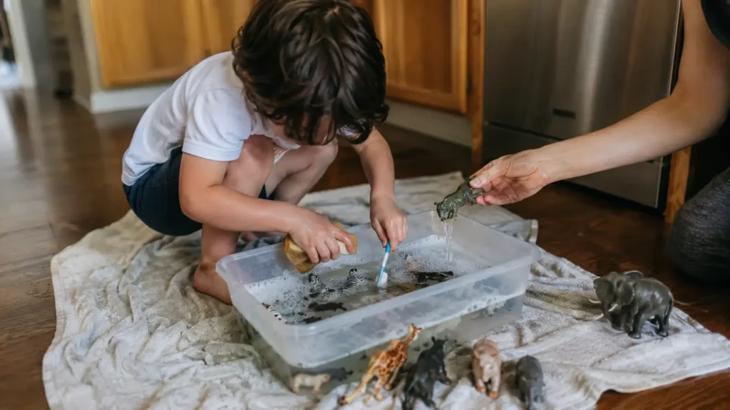 A boy kneels on a kitchen floor washing plastic safari animals in a bowl of water with a towel beside him