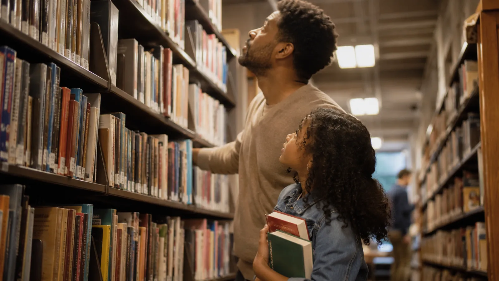A dad and his daughter look up at tall library shelves, choosing books together
