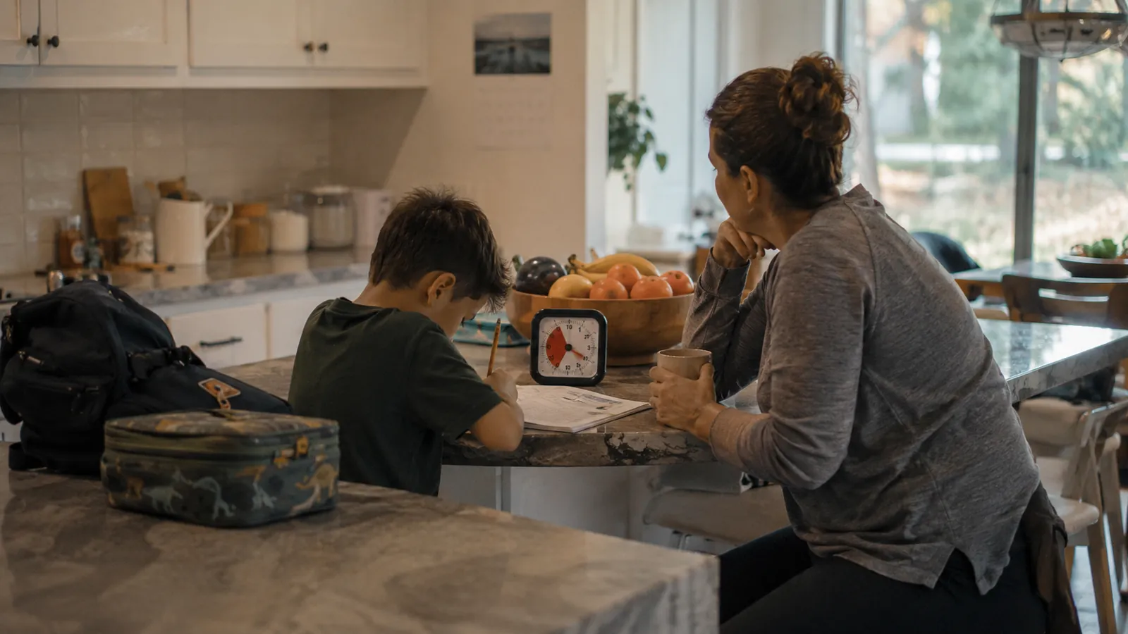 A boy works at a kitchen island with a visual timer while his mom sits across from him