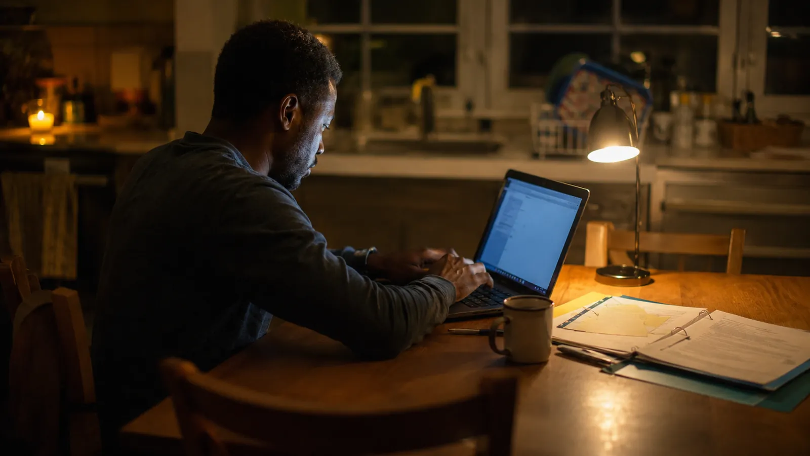A dad sits alone at a table late at night, lit by a laptop screen and a small lamp