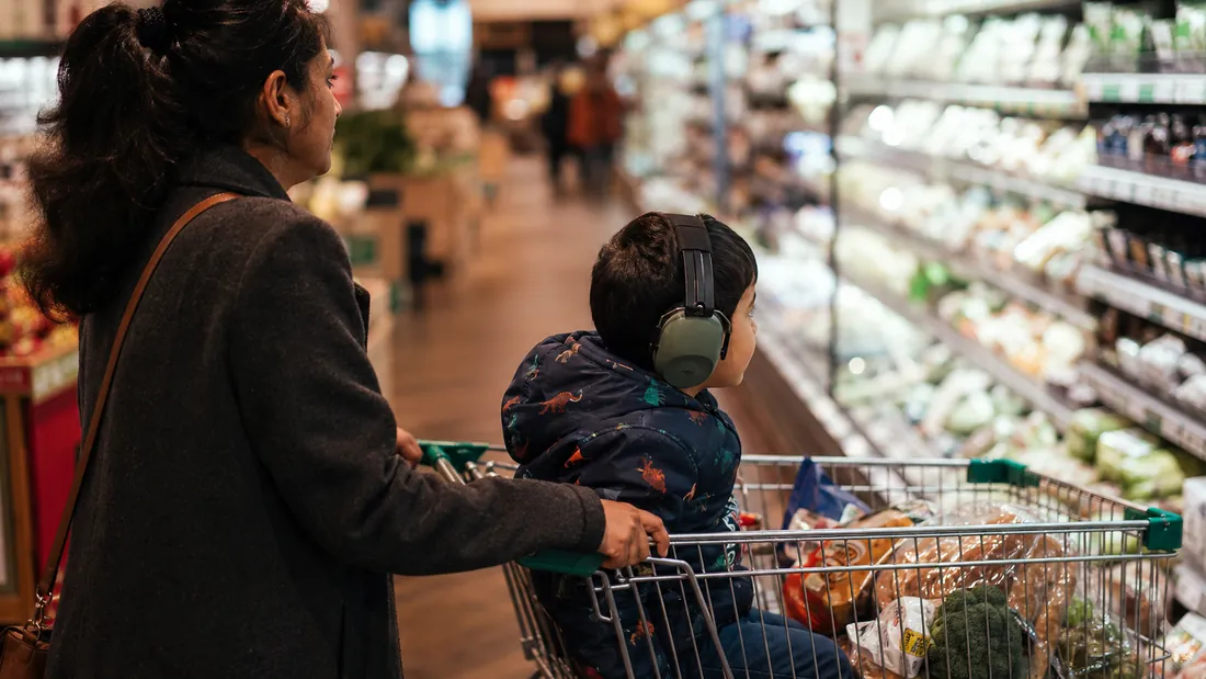 A child in ear defenders rides in a shopping cart down a bright supermarket aisle