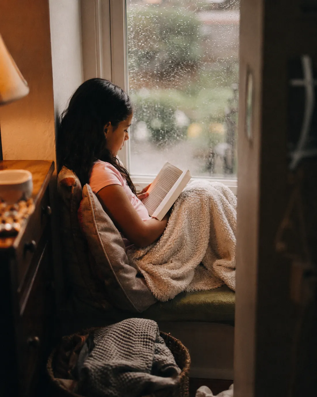 A child curls up with a book in a small reading nook by the window