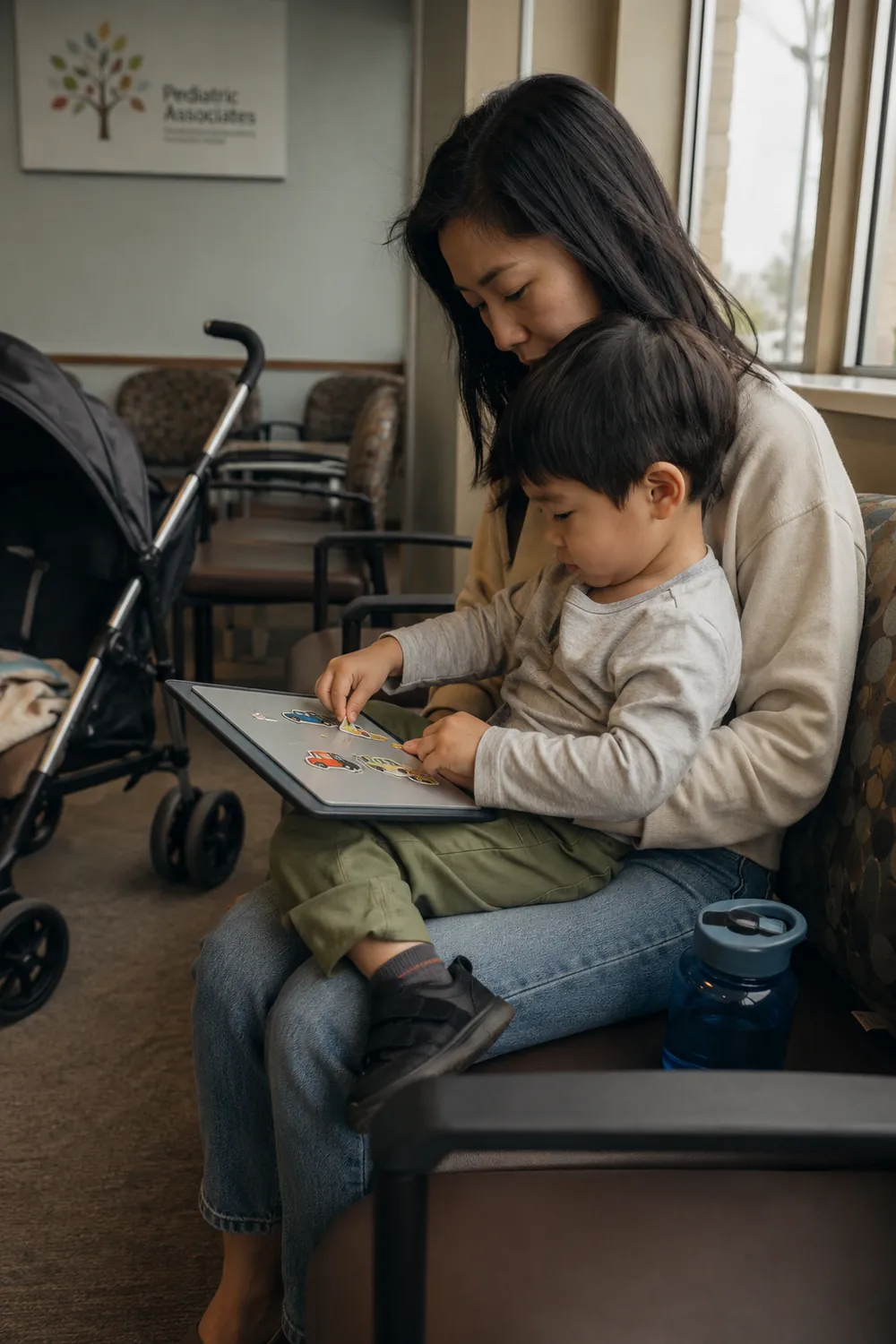 A mother sits with her toddler son on her lap in a pediatric office waiting room while he plays on a tablet