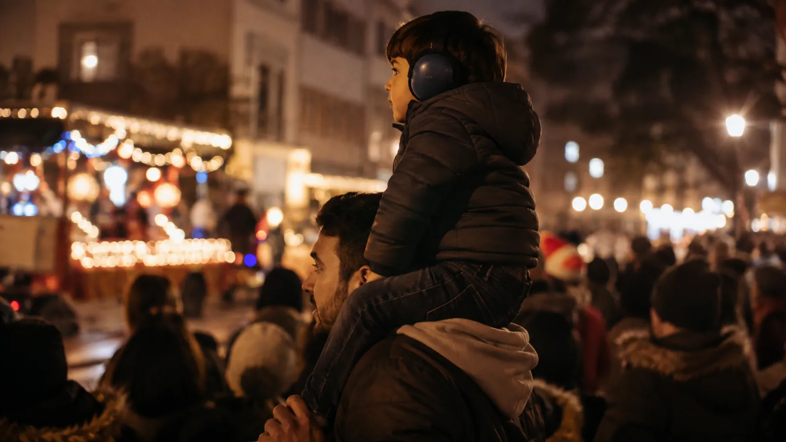 A child in ear defenders sits on a parent's shoulders in a crowd at a lit-up evening festival