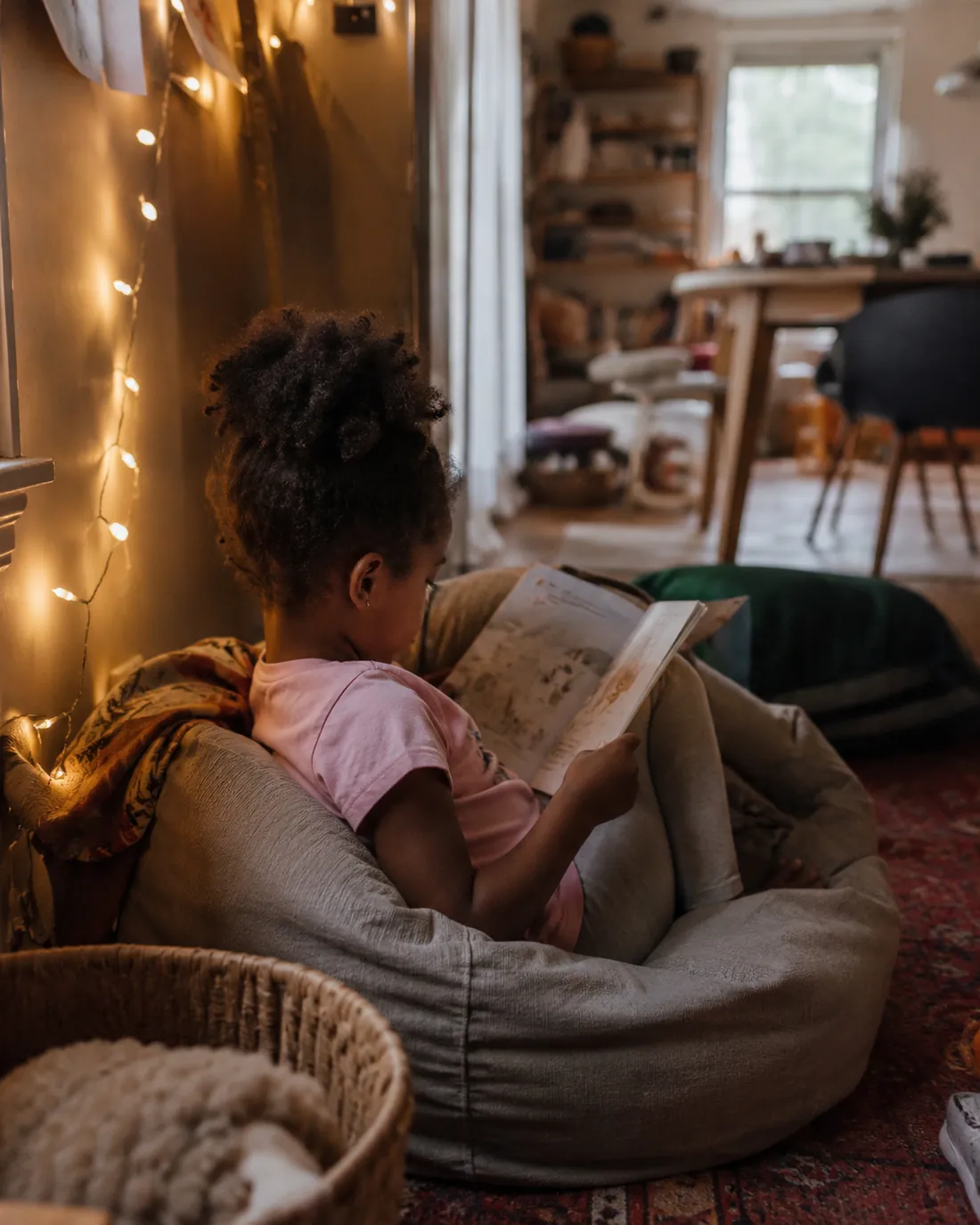 A girl reads in a quiet corner on a floor cushion under fairy lights