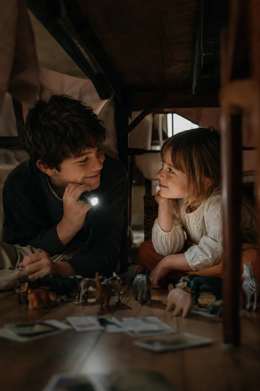 Two children lie under a table lit only by a flashlight, surrounded by small toy animal figures