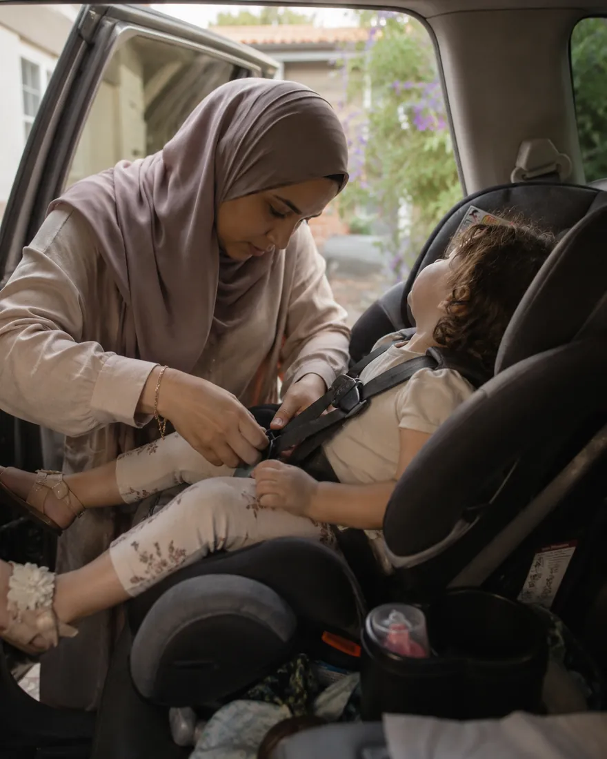 A mother buckles a resisting toddler into a car seat through the open back door before a road trip