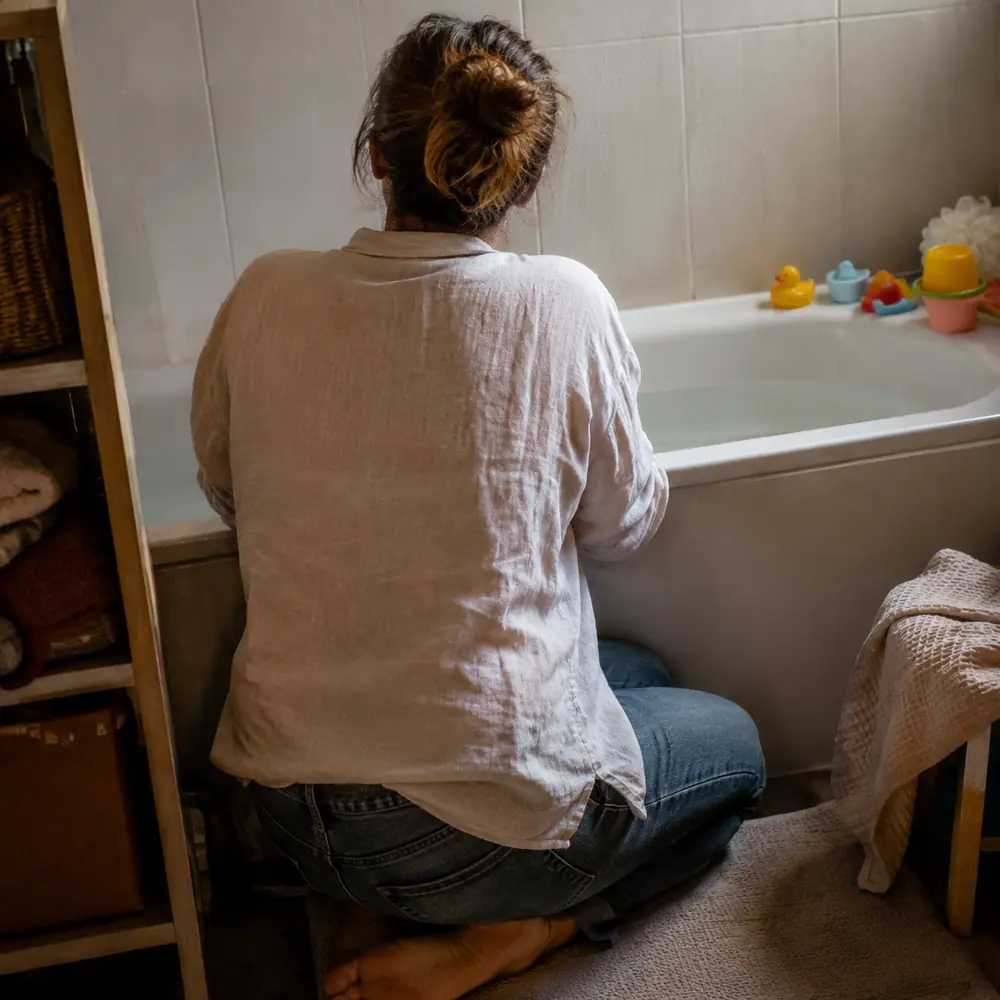 A parent kneeling beside a bath tub with a towel over one arm