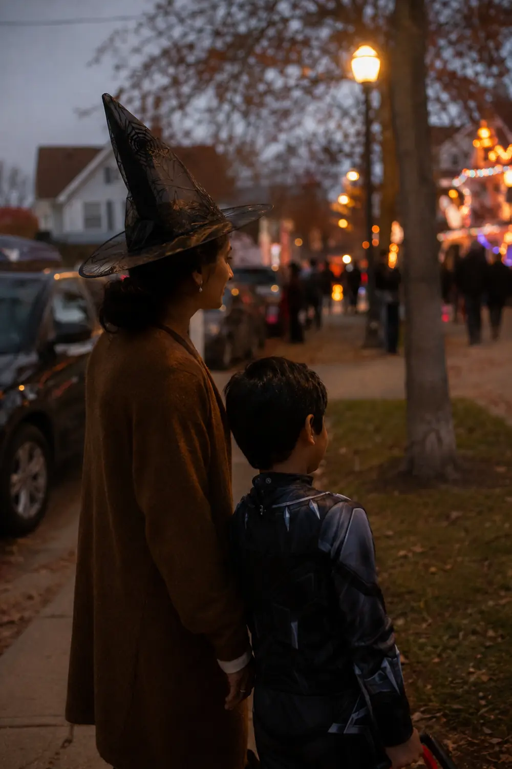 A mom in a witch hat and a young boy on a sidewalk at night looking toward decorated houses from a distance