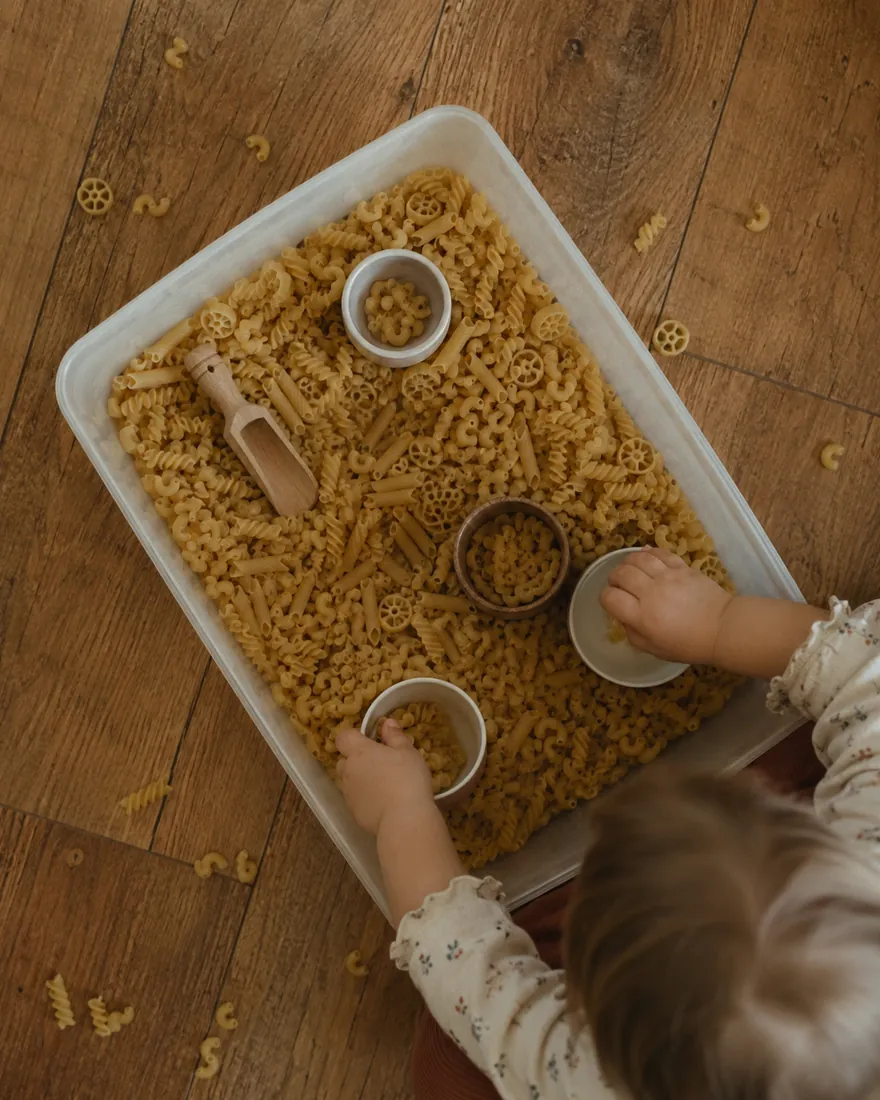 Overhead view of a toddler scooping dry pasta into small bowls inside a plastic bin on a wooden floor
