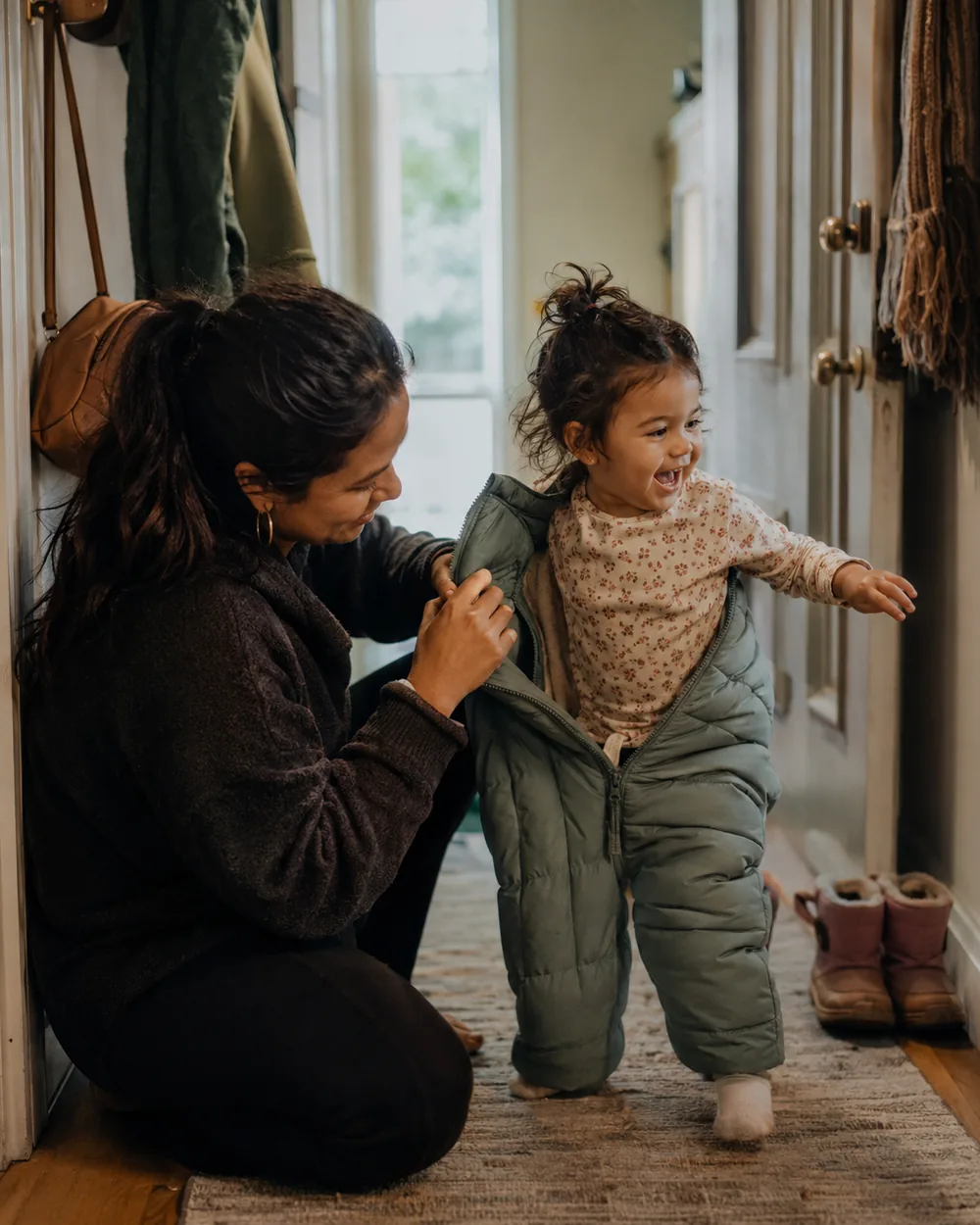 A toddler wriggling out of a snowsuit in a hallway while a parent holds a sleeve