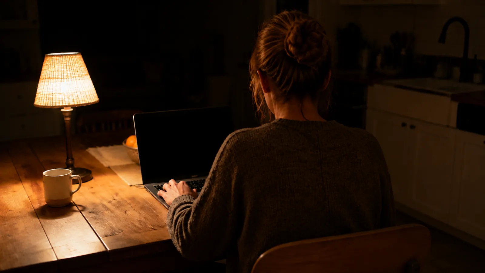 Adult seen from behind typing on a laptop at a wooden kitchen table, one lamp lit and the rest of the house dark